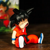 A small anime figurine of a child Goku in a sitting pose, wearing his classic red and black gi with blue and white shoes, set on a dark wooden surface.