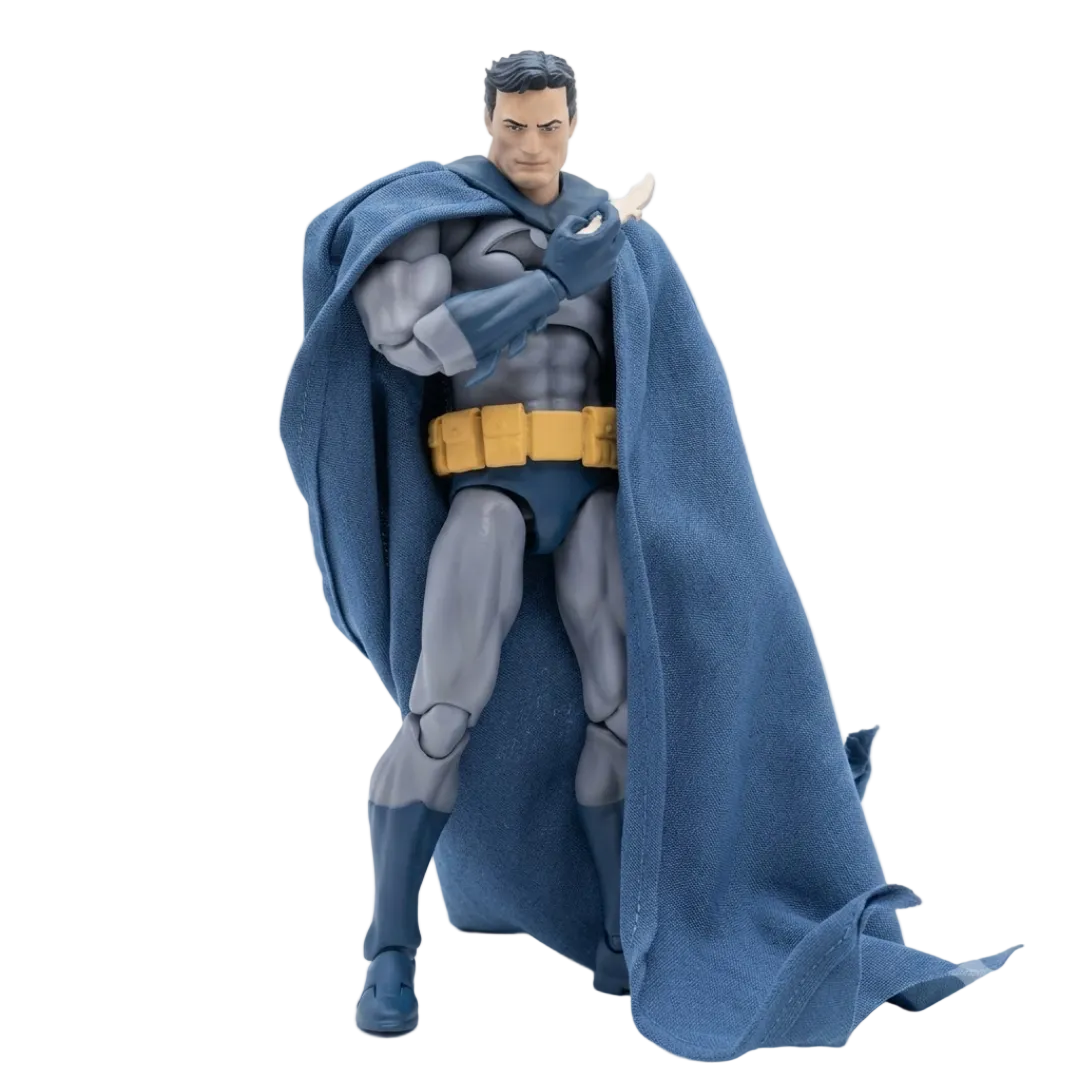 A Batman action figure posed with one arm raised, wearing a textured blue cape, gray and blue bodysuit, and yellow utility belt, photographed against a clean white studio background.