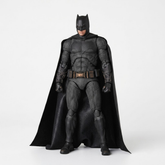 A Batman action figure or collectible statue in a full-body pose, wearing a textured black suit with a flowing black cape, standing against a plain light gray studio background.
