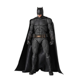 A Batman action figure or collectible statue in a full-body pose, wearing a textured black suit with a flowing black cape, standing against a plain light gray studio background.