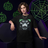 A model wearing a black graphic t-shirt featuring a metallic robot face design with green accents, posed against a dark background with neon green pumpkin and spiderweb graphics.