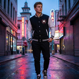 A man wearing a black and white varsity jacket with a spider graphic on the chest, black sweatpants, and sneakers, standing on a wet city street at dusk.