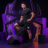 A man modeling black athletic wear, including a fitted short-sleeve t-shirt and matching shorts with white spider-themed graphics, posed in front of a stylized purple and black geometric set with spiderweb motifs.