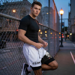 A man leans against a chain-link fence at dusk, wearing a fitted black short-sleeve athletic shirt and white drawstring shorts with black side panels and white text near the hem.