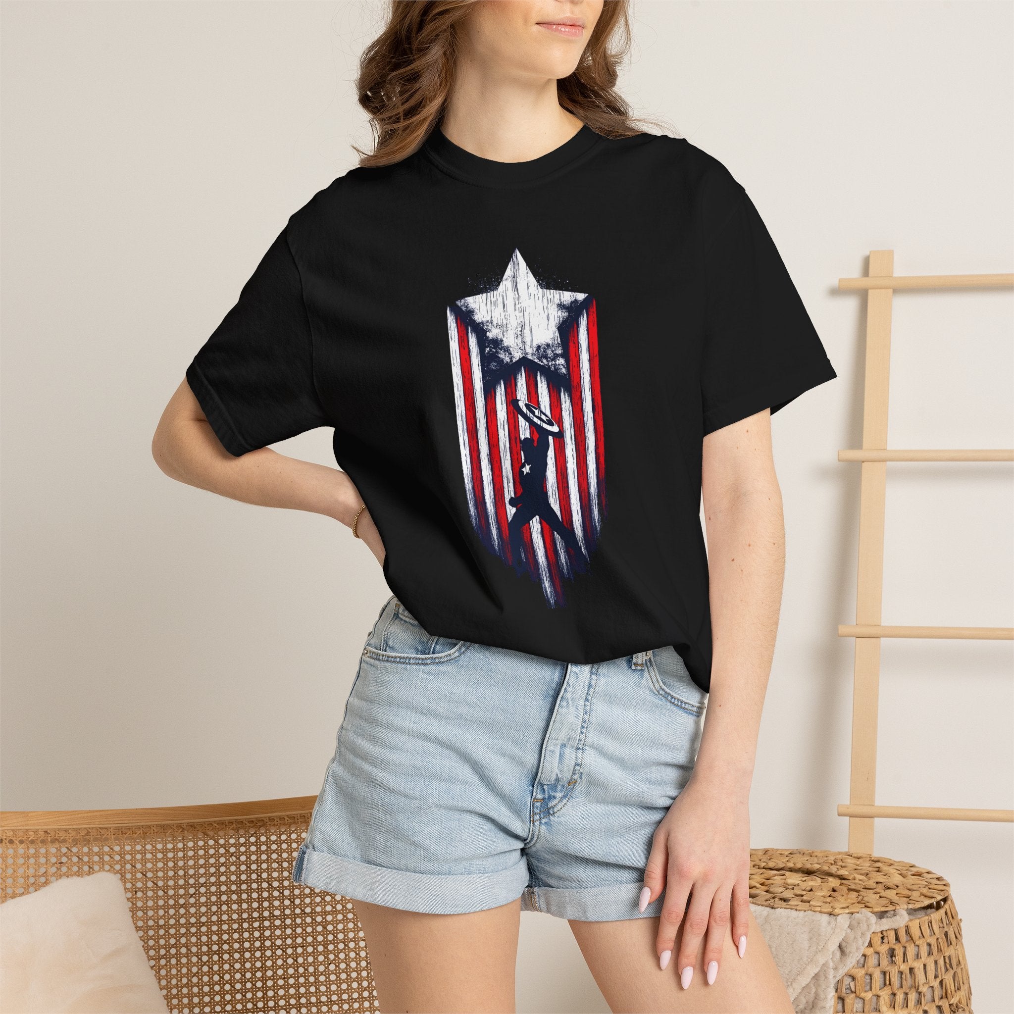 A woman wearing a black short-sleeve t-shirt with a distressed red, white, and blue shield graphic on the front, paired with light-wash denim shorts in a neutral indoor setting.