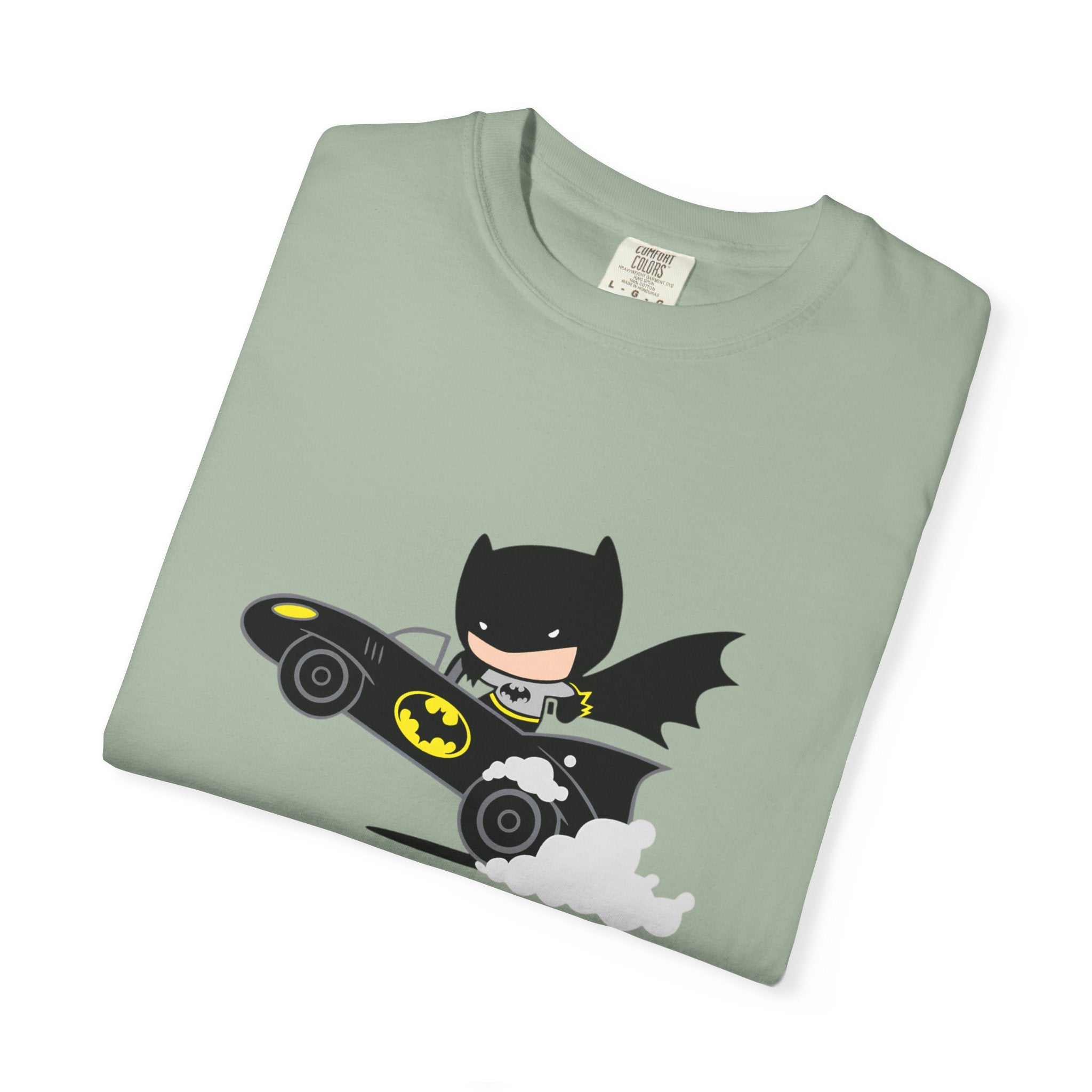 A folded sage green crewneck t-shirt featuring a cartoon graphic of Batman riding his black and yellow Batmobile with white smoke, displayed on a clean white background.