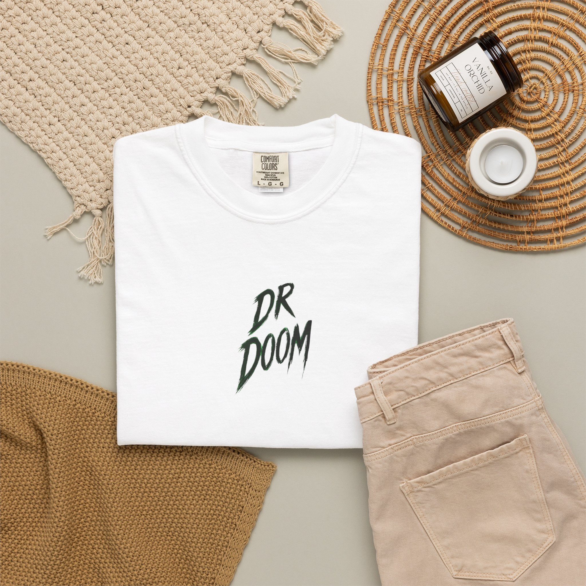 A folded white T-shirt with a small mountain landscape graphic on the chest, styled in a flat lay with beige pants, a woven blanket, a candle jar, and a small cup on a neutral background.