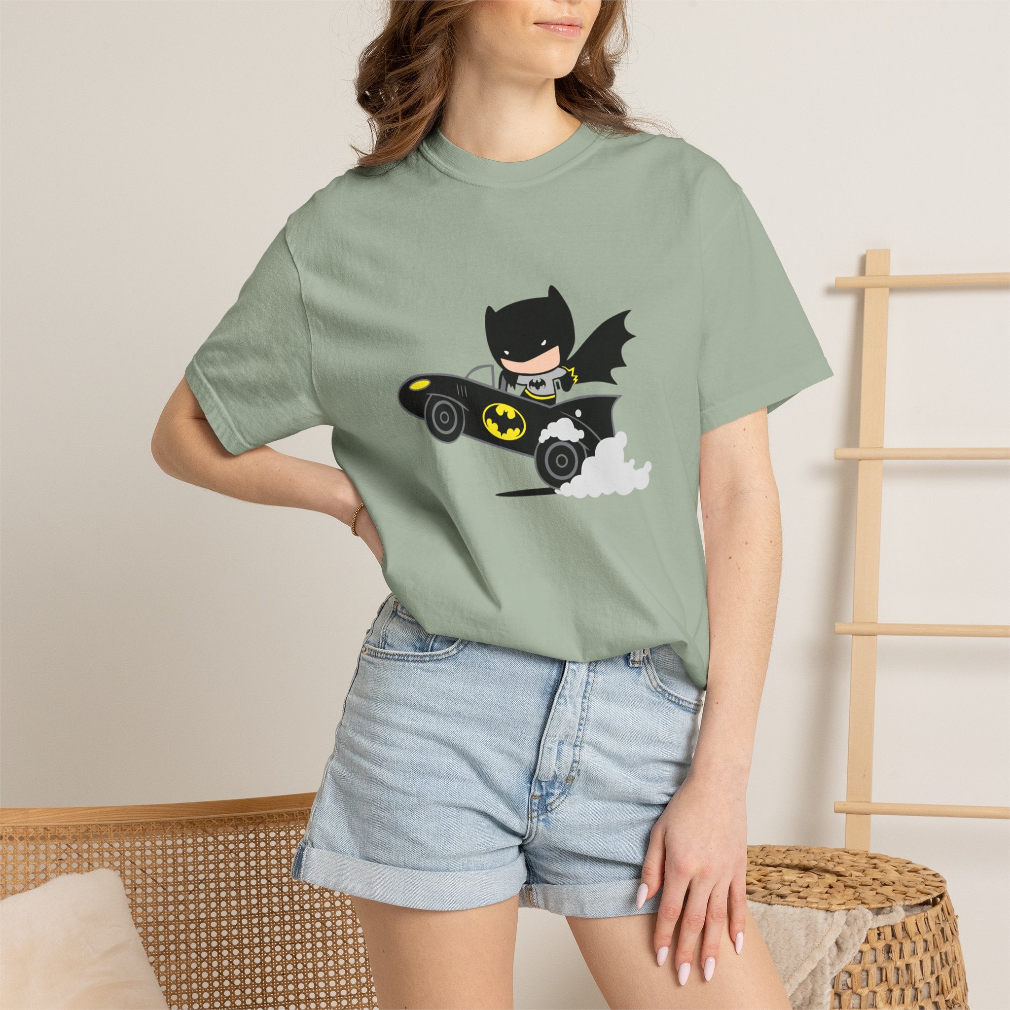 A woman wearing a sage green graphic t-shirt with a cartoon Batman driving a black and yellow car, paired with light-wash denim shorts in a cozy indoor setting.