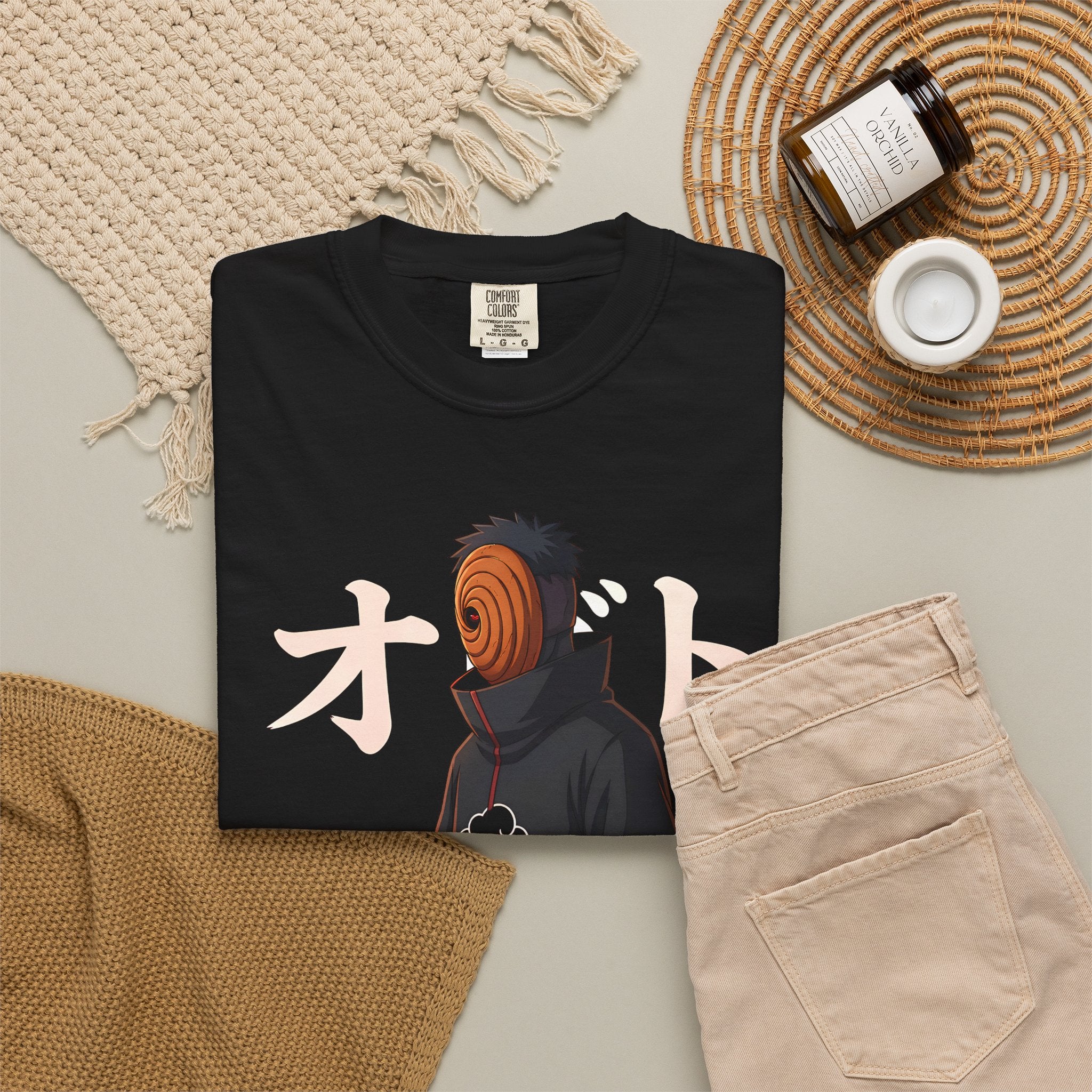 A folded black graphic T-shirt is arranged in a styled flat lay with beige pants, cream knit textiles, a woven placemat, a small jar candle, and a white cup on a light gray surface. The shirt graphic shows a dark character with an orange spiral mask and large white Japanese characters.