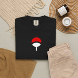 A folded black T-shirt with a red and white leaf emblem on the chest, styled in a flat lay with beige pants, a woven placemat, a candle jar, and a small cup on a neutral background.