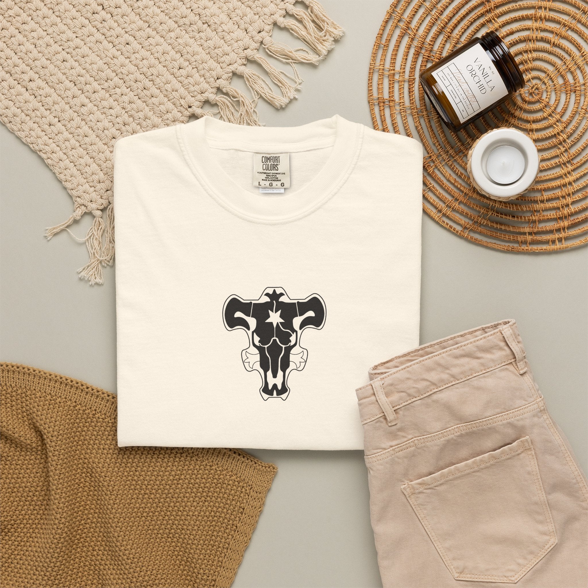 A folded cream-colored t-shirt with a black bull skull graphic on the chest, styled in a flat lay with beige pants, a woven blanket, a candle jar, and a small cup on a neutral background.
