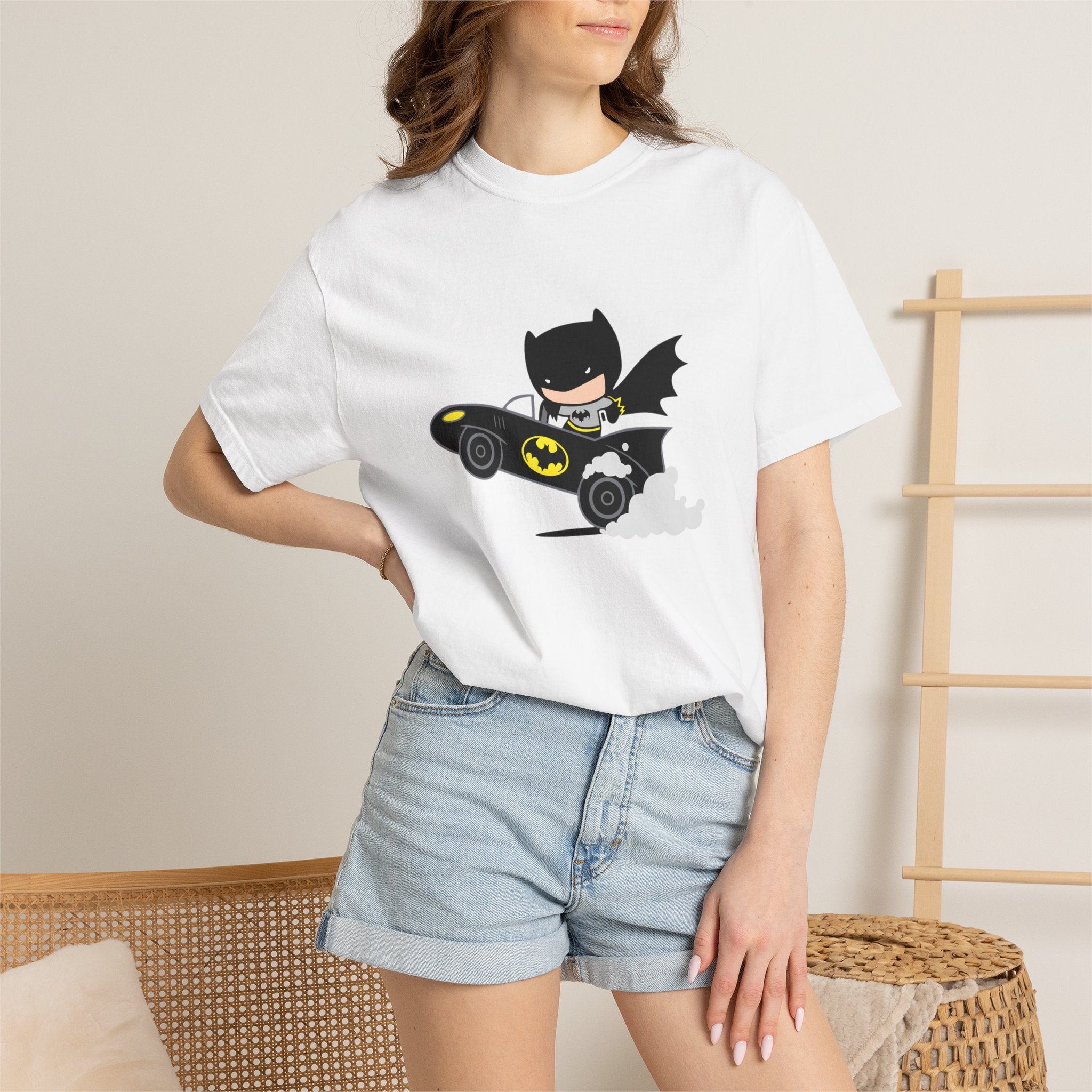 A woman wearing a white short-sleeve graphic T-shirt with a cartoon Batman driving a black and yellow Batmobile, paired with light blue denim shorts in a softly lit indoor setting.