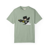 A sage green short-sleeve crewneck t-shirt featuring a cartoon graphic of Batman driving a black Batmobile with yellow accents and white smoke, displayed on a clean white background.