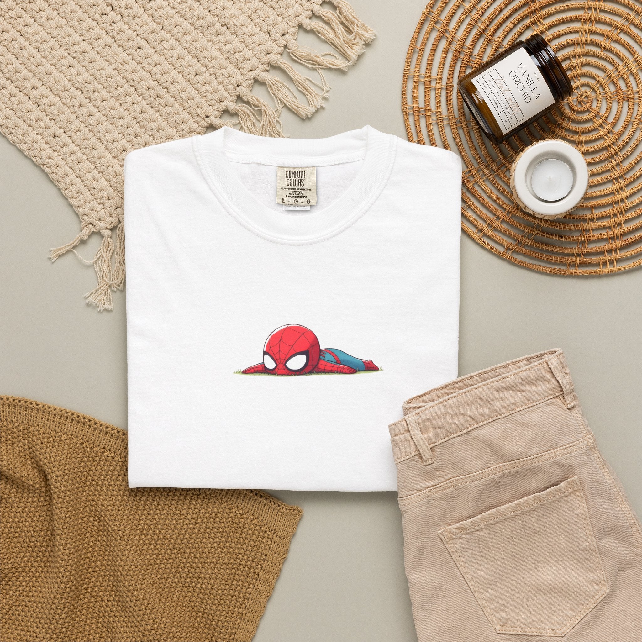A folded white t-shirt featuring a small graphic of a red and blue Spider-Man head peeking out from behind a grassy patch, styled in a flat lay with beige pants, a woven blanket, a candle, and a jar on a neutral background.