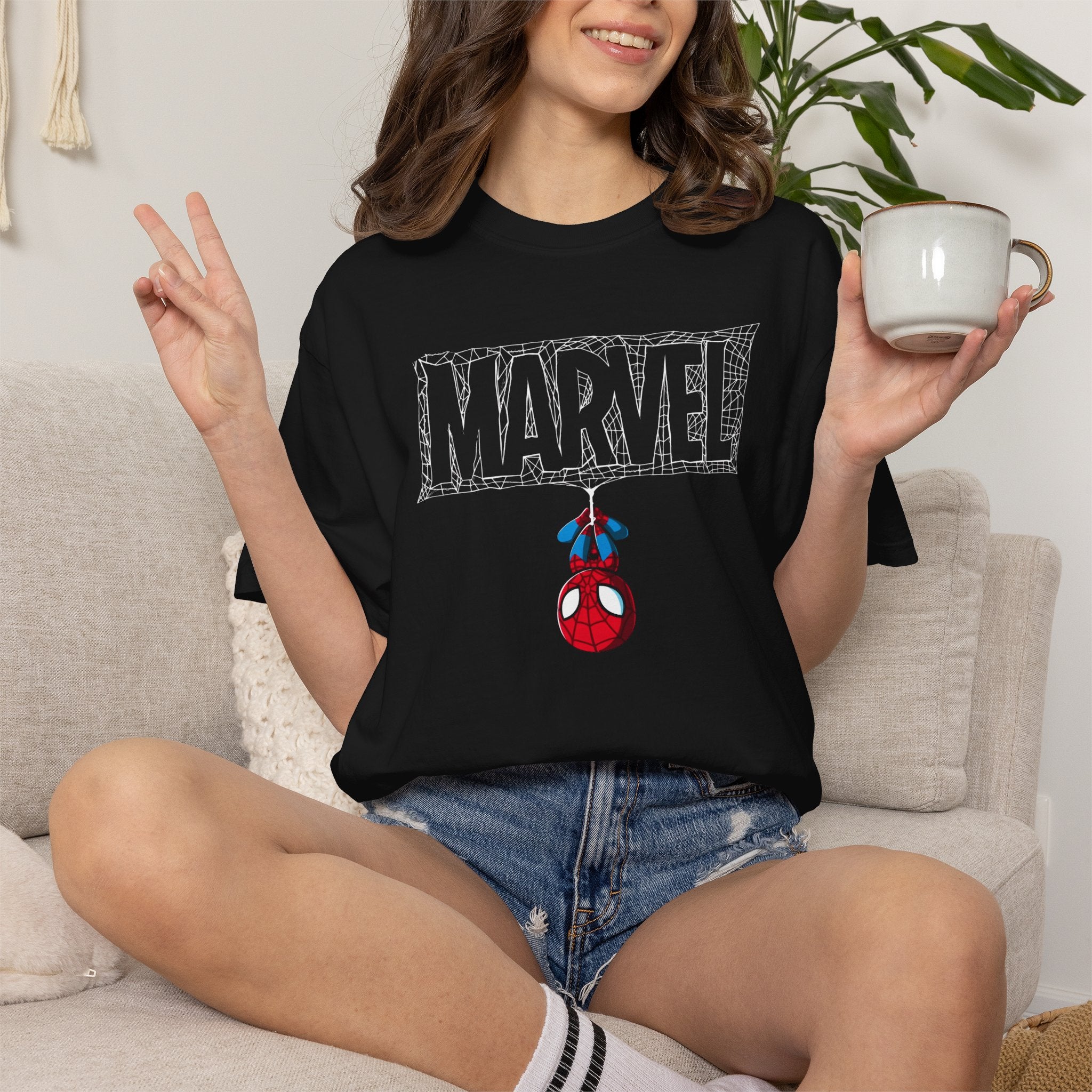 A woman sitting on a light beige couch wearing a black graphic t-shirt with a 'MARVEL' webline design and Spider-Man hanging from it. She is holding a white ceramic mug.