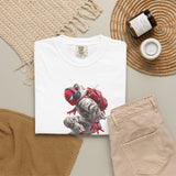 A folded white T-shirt with a graphic print of a cat dressed as a superhero in red and gray, styled in a flat lay with beige pants, a woven blanket, a candle, and a small cup on a neutral background.