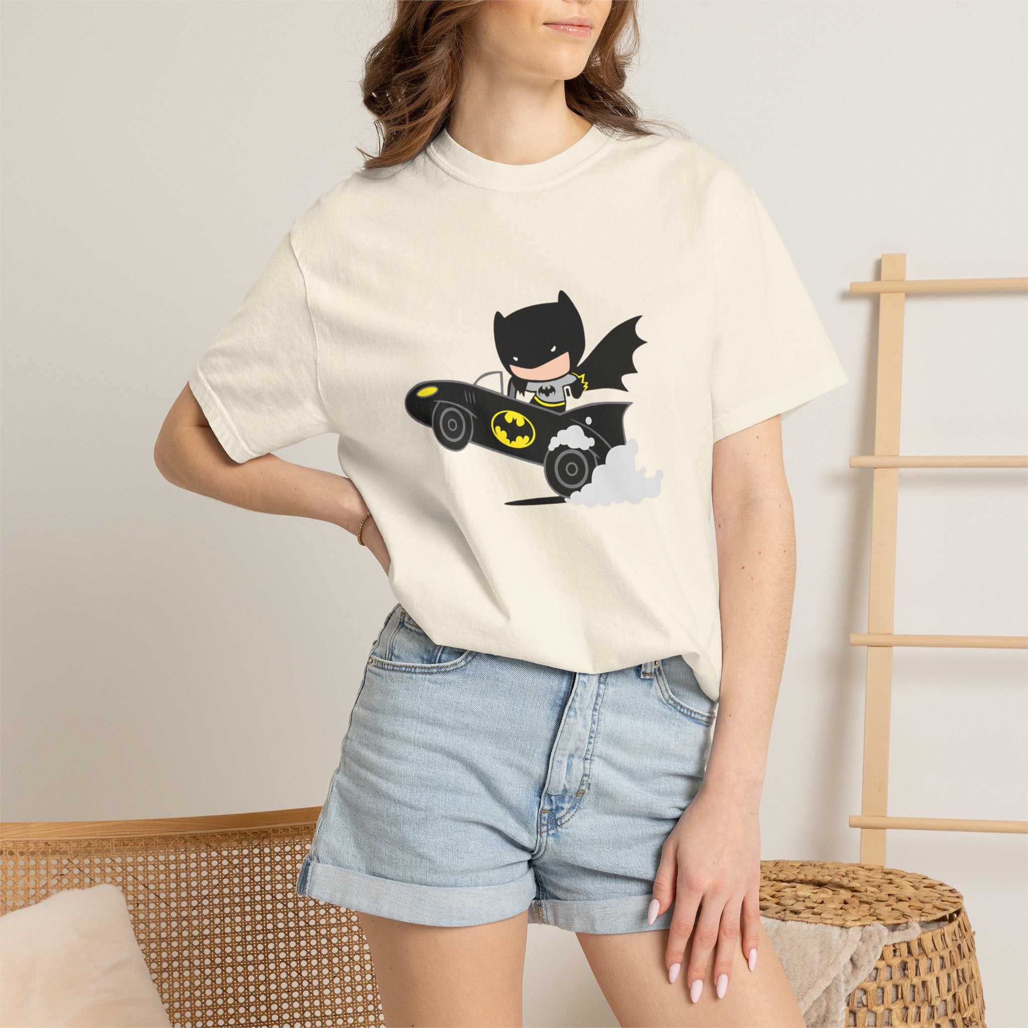 A woman wearing a cream-colored t-shirt with a cartoon graphic of Batman driving a black Batmobile, paired with light-wash denim shorts in a cozy indoor setting.
