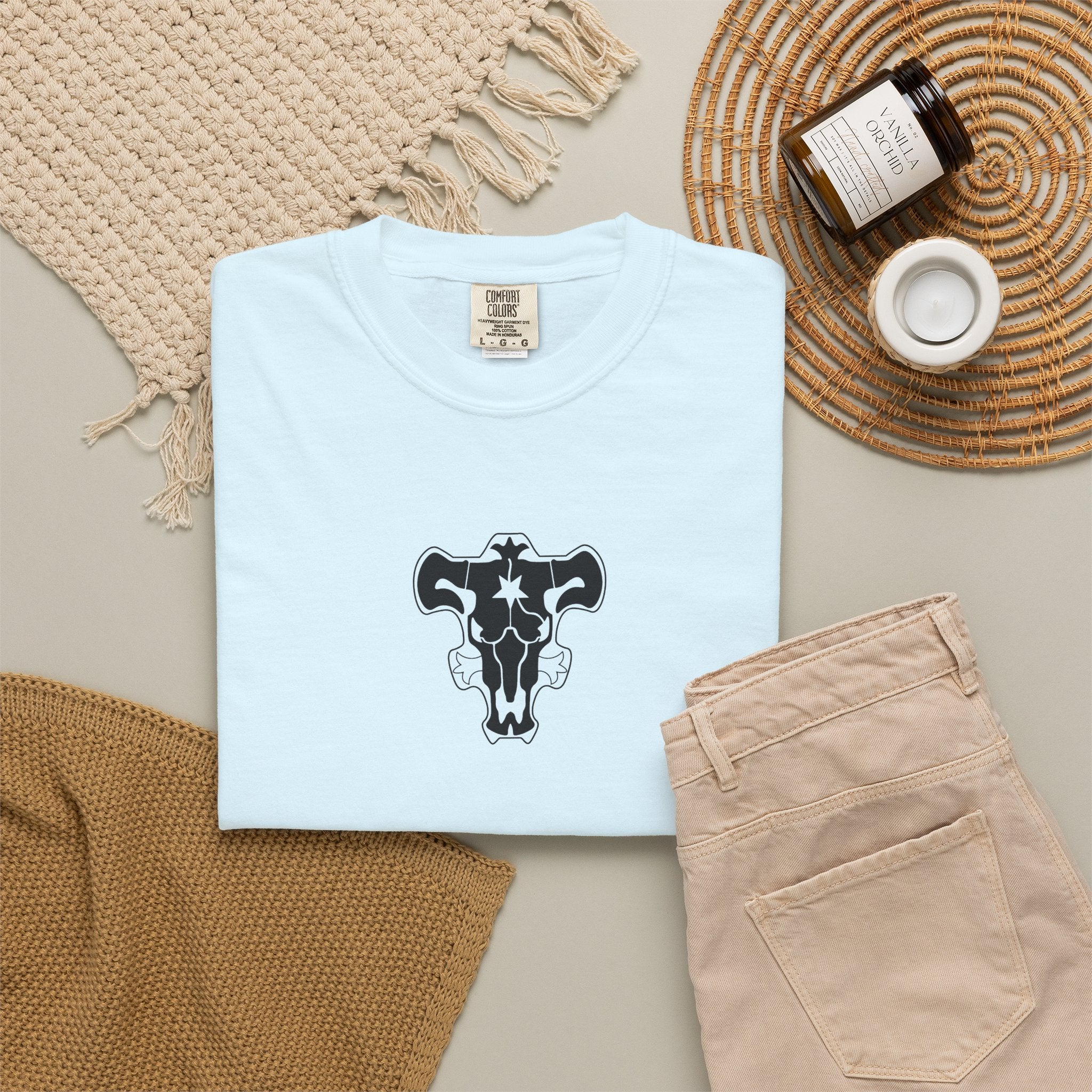 A folded light blue t-shirt with a black and white bull skull graphic on the chest, styled in a flat lay with beige pants, a woven blanket, a candle, and a jar of beeswax candles on a neutral background.