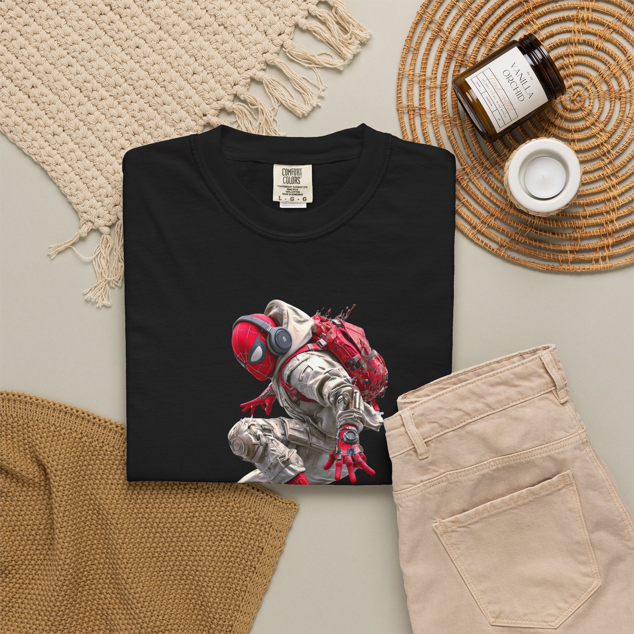A folded black T-shirt with a graphic print of a red and white superhero crouching, styled in a flat lay with beige pants, a woven placemat, a candle jar, and a knit blanket on a light gray surface.