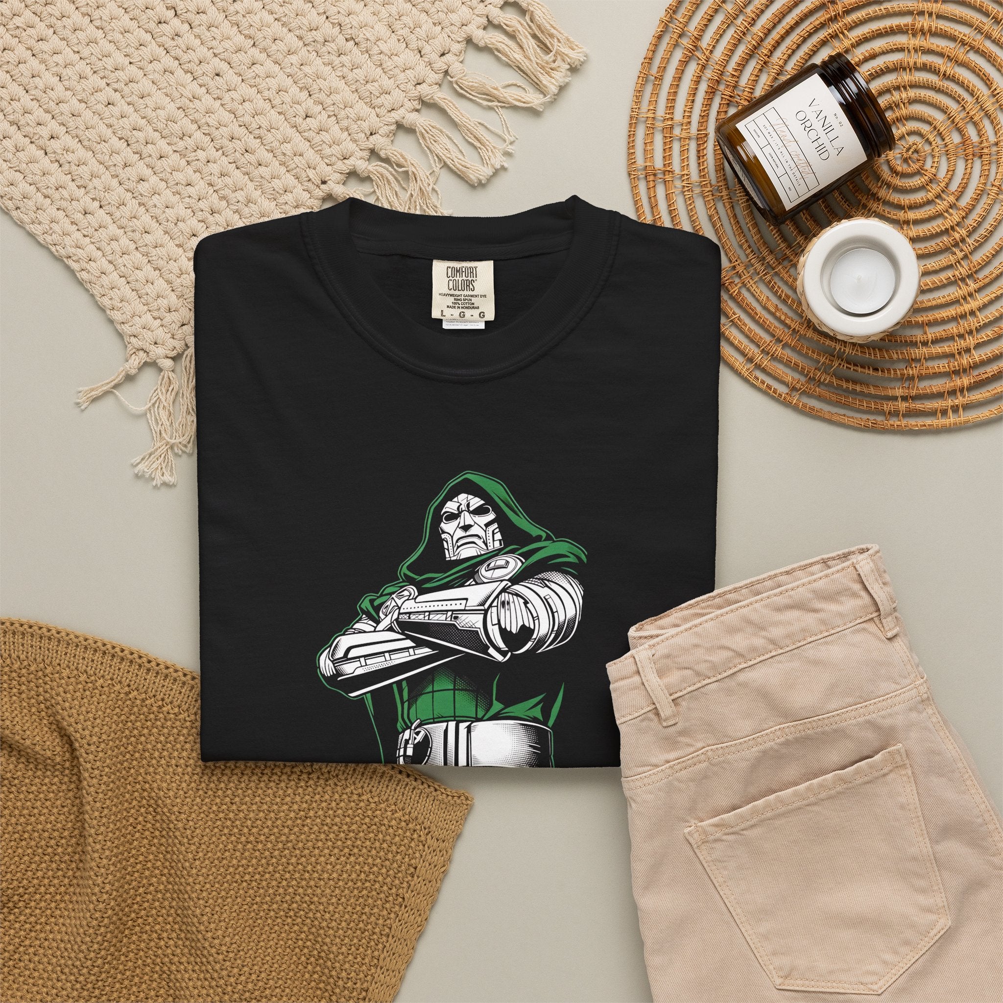 A folded black graphic T-shirt is arranged in a styled flat lay with beige pants, a woven placemat, a small candle jar, and a cream knit textile on a light neutral background. The shirt features a printed comic-style character in green, white, and gray with crossed arms.