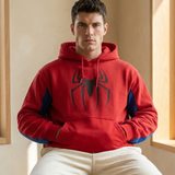 A man wearing a red Spider-Man themed hoodie with a large black spider logo on the chest and blue accents on the sleeves, paired with cream sweatpants, sitting in a warm indoor setting.