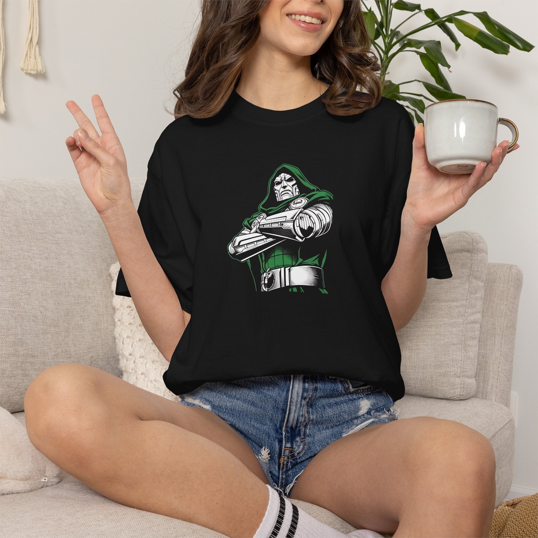 A woman sitting on a light beige couch wearing a black graphic t-shirt with a green and white Robotech character print. She is holding a white ceramic mug in one hand and making a peace sign with the other, with a houseplant in the background.