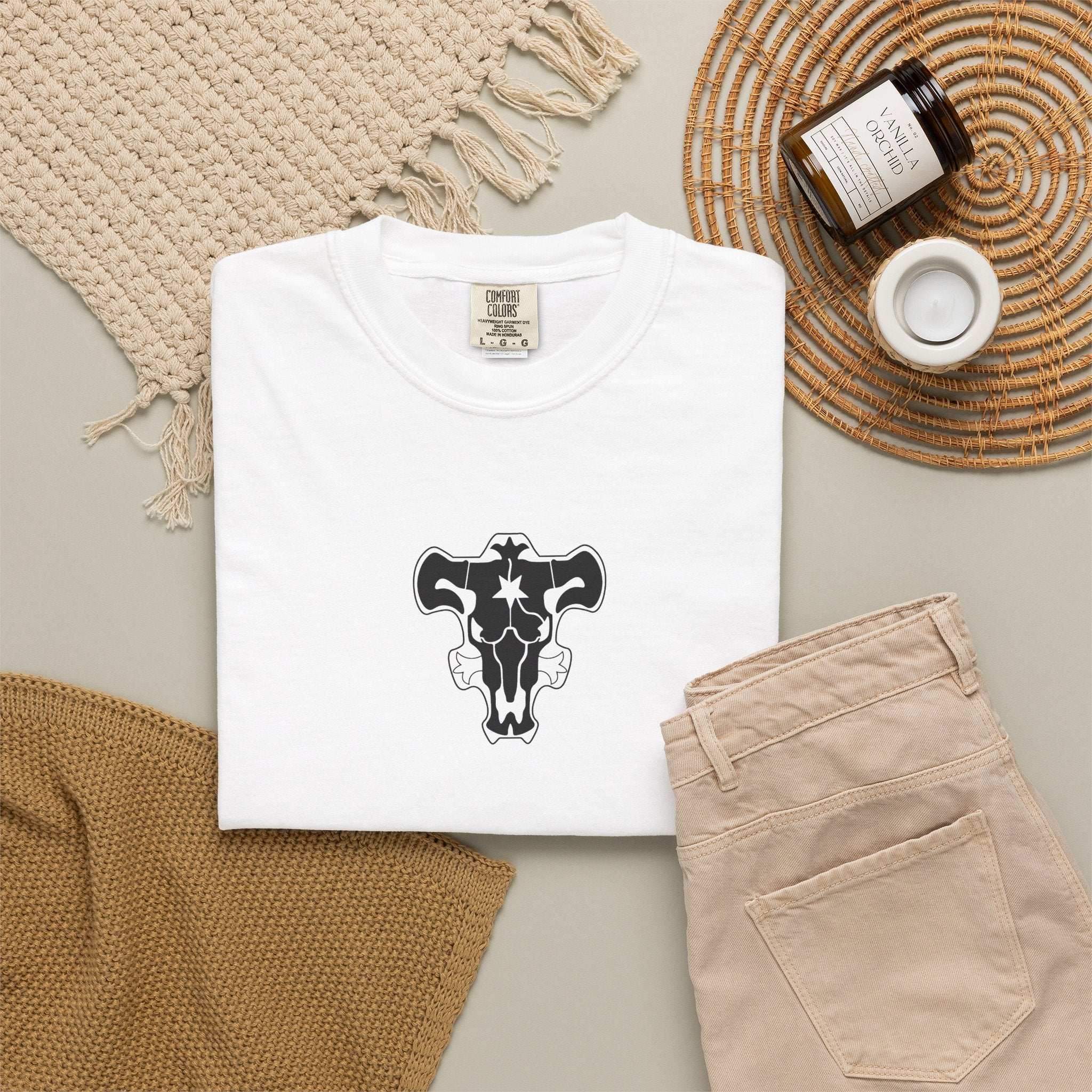 White t-shirt with a graphic design on a neutral surface with beige pants and decorative items.