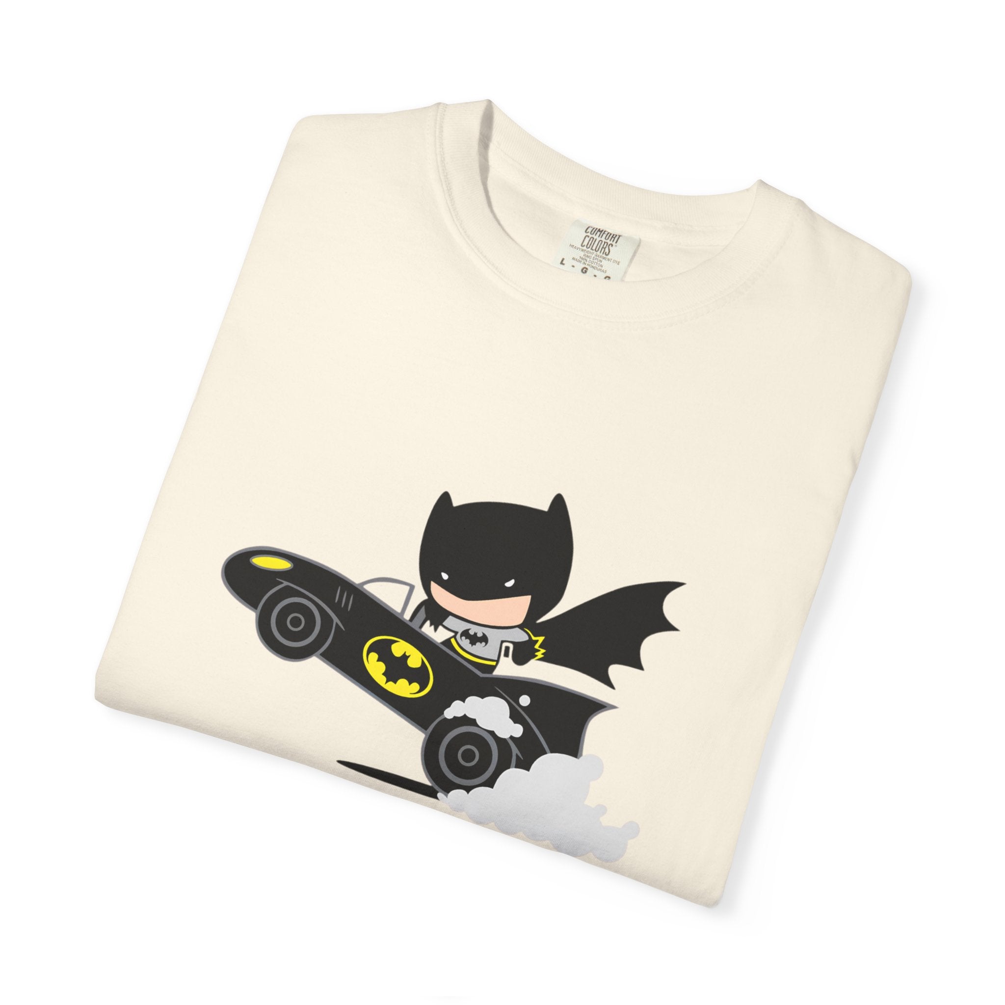 A folded cream-colored t-shirt featuring a cartoon graphic of Batman riding his black Bat-bat car with yellow accents and white smoke, displayed on a clean white background.