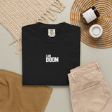 A folded black T-shirt with the text 'I AM DOOM' in white block letters is arranged in a styled flat lay on a light gray surface, surrounded by beige knit textiles, folded beige pants, a small jar candle, and a white cup.