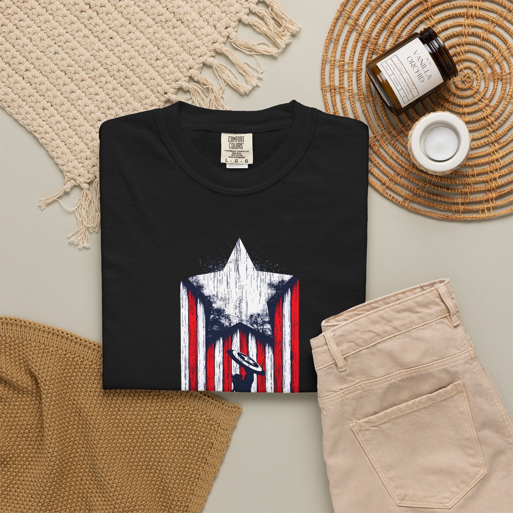 A folded black T-shirt with a distressed red, white, and blue star-and-stripes graphic on the front, styled in a flat lay with beige pants, a woven placemat, a candle jar, and a small cup on a light neutral background.
