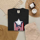 A folded black T-shirt with a distressed red, white, and blue star-and-stripes graphic on the front, styled in a flat lay with beige pants, a woven placemat, a candle jar, and a small cup on a light neutral background.