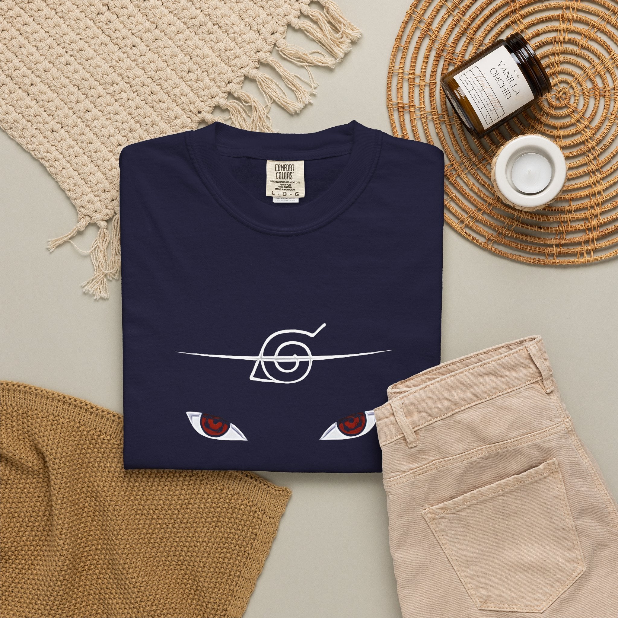 A navy blue folded t-shirt with a white line art graphic of a sloth wearing a chef's hat and stirring a pot, styled in a flat lay with beige pants, a woven blanket, a candle, and a jar on a neutral background.