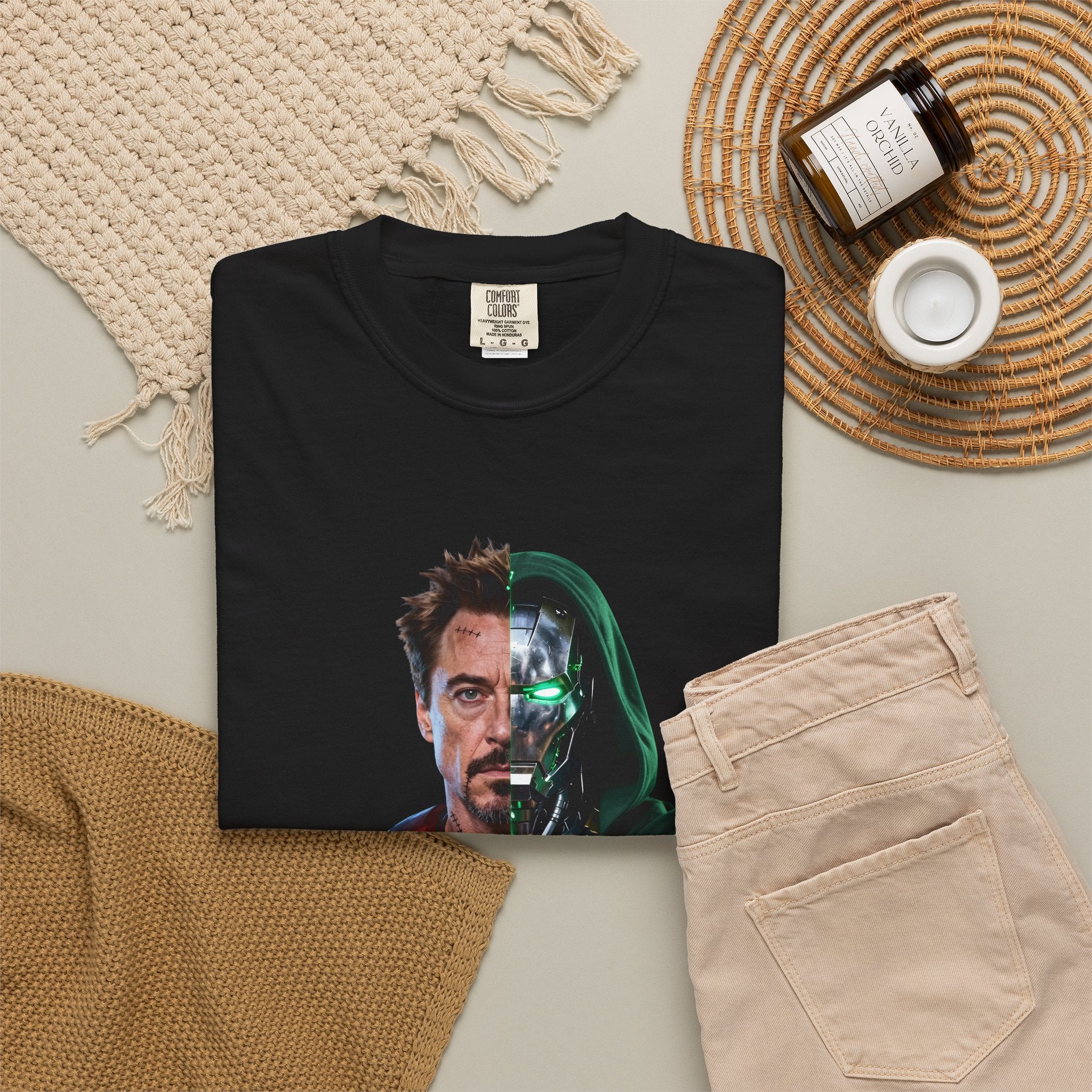 A folded black T-shirt is displayed in a styled flat lay on a light beige surface. The shirt features a split-face graphic: one half shows a man's face, and the other half shows a metallic robotic face with glowing green eyes.