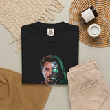 A folded black T-shirt is displayed in a styled flat lay on a light beige surface. The shirt features a split-face graphic: one half shows a man's face, and the other half shows a metallic robotic face with glowing green eyes.
