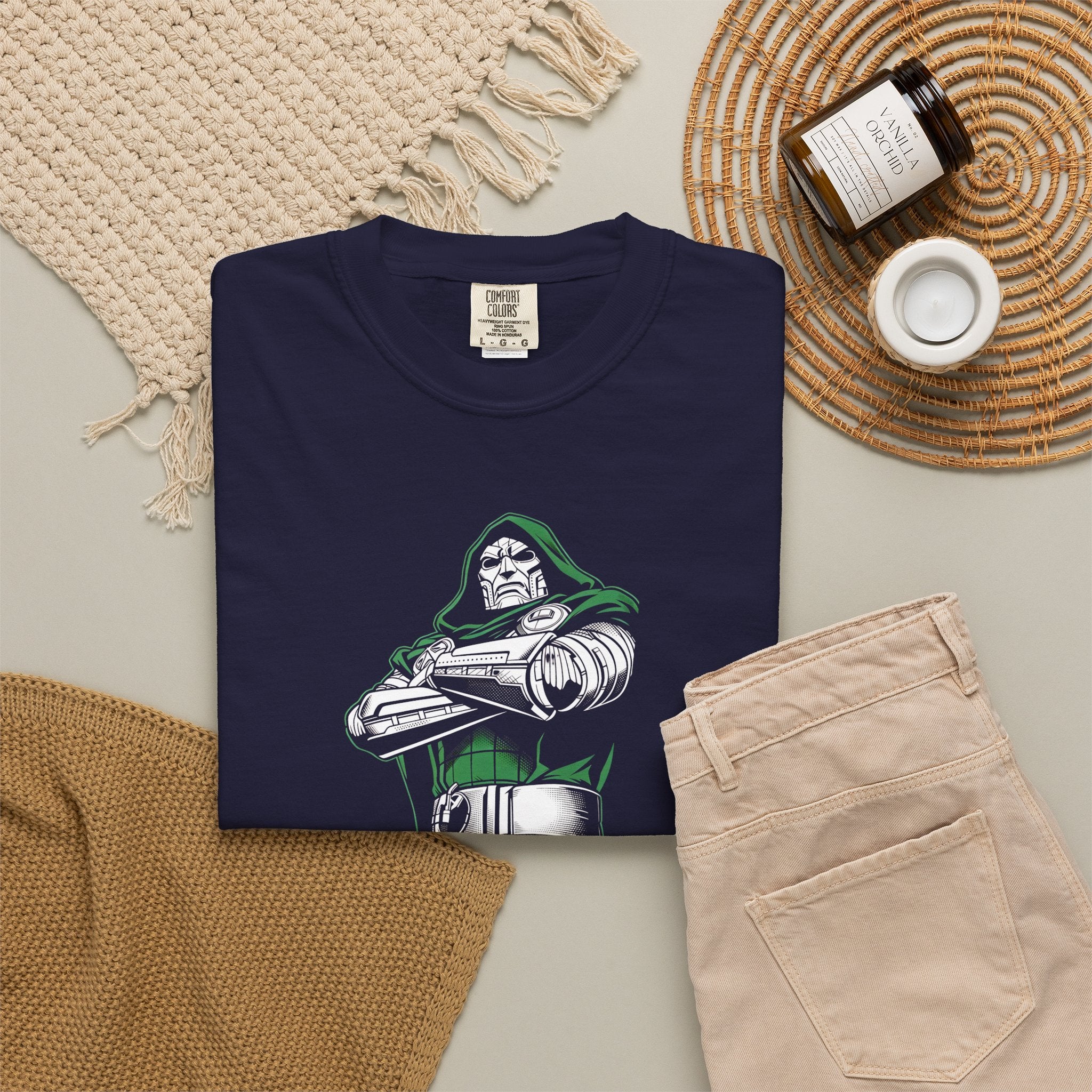 A folded navy blue t-shirt with a graphic of a green-robed skeleton character, styled in a flat lay with beige pants, a woven blanket, a candle, and a small cup on a neutral background.