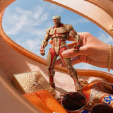 A hand holds a muscular anime action figure with exposed muscle detailing, posed inside a beige leather bag alongside sunglasses, a gold compact, and a patterned scarf, with a blue sky visible through the bag's opening.