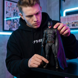 A man in a black hoodie holds a detailed Batman action figure with a dark cape and textured suit, set against a modern room background with blue lighting.