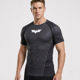 A man wearing a dark gray and black textured athletic compression shirt with a white Batman logo on the chest, photographed against a light gray studio background.