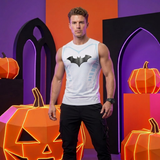 A man stands in a studio setting wearing a white sleeveless tank top with a black bat logo on the chest, paired with black pants. The background features Halloween-themed props including orange jack-o'-lanterns and purple gothic arches.