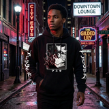 A man wearing a black pullover hoodie with a large anime-style graphic on the chest and white text down the sleeves, standing on a wet city street at night with neon signs in the background.