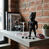 A Black Panther Funko Pop! vinyl figure stands on a concrete shelf in front of a brick wall, with its clear stand, alternate hands, and Captain America shield nearby. Books, a camera, and a small plant are arranged around it in a styled indoor setting.