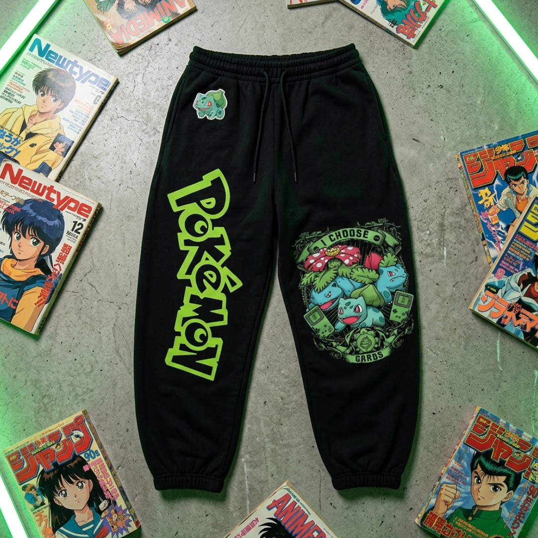 Black jogger sweatpants featuring a large neon green 'Pokémon' logo on the left leg and a colorful 'I CHOOSE BULBLEEP' graphic on the right leg, displayed flat on a gray concrete surface surrounded by vintage anime magazines.