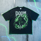 A black short-sleeve graphic t-shirt featuring the word 'DOOM' in distressed white letters above a green and black comic-style character illustration, laid flat on a gray concrete-textured background with neon green and blue light effects.