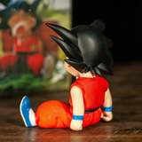 Toy figure of a character in a red outfit with black spiky hair on a wooden surface, Dragon Ball Super logo in the background.