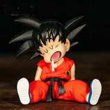 A collectible figurine of a baby Goku from Dragon Ball Z, sitting down with his signature black spiky hair, red gi, and black belt. The figurine is posed with its mouth open, showing a white tooth, set against a dark background.