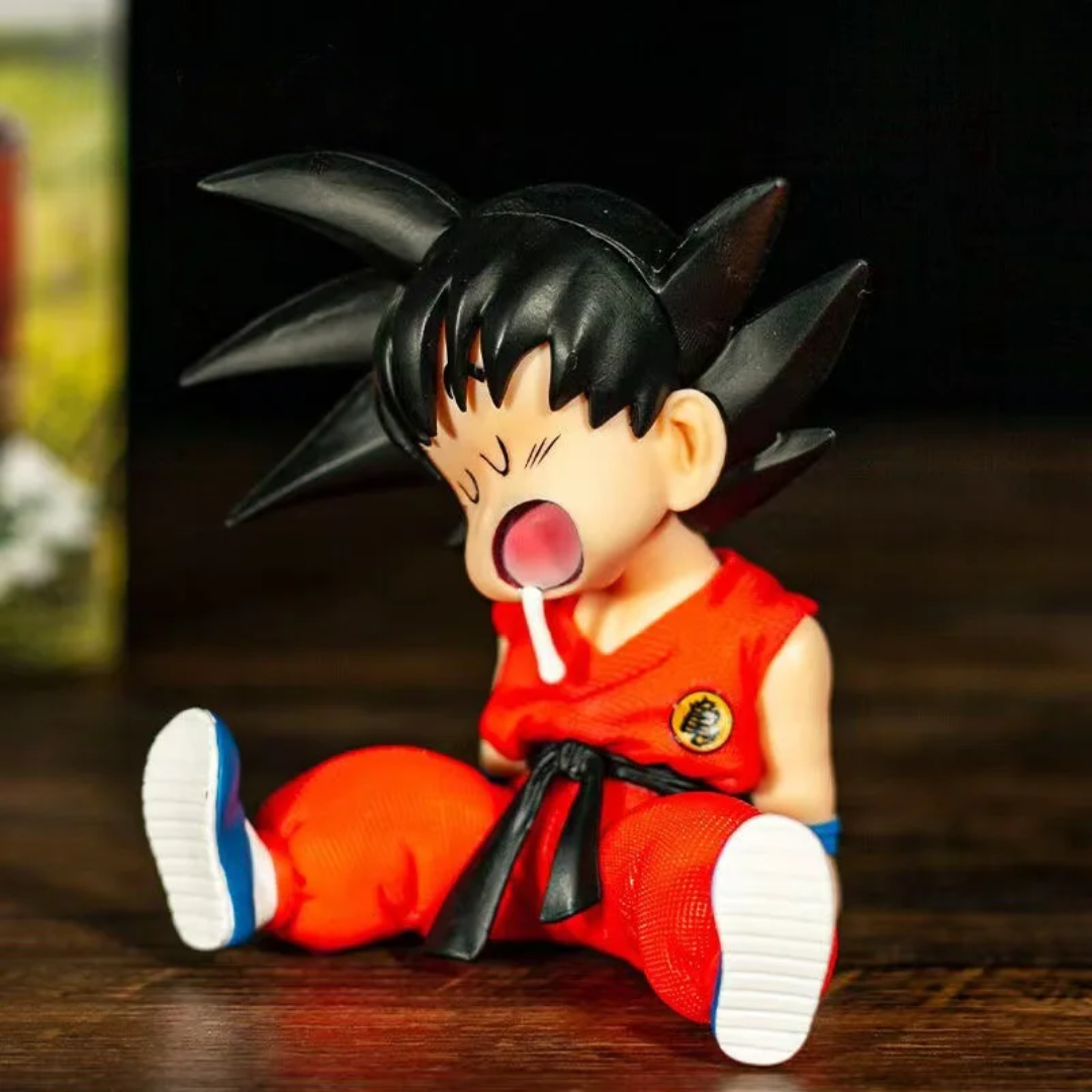 A small anime figurine of a child Goku sitting on a dark wooden surface, wearing his signature red and black outfit with white and blue shoes, against a dark background.