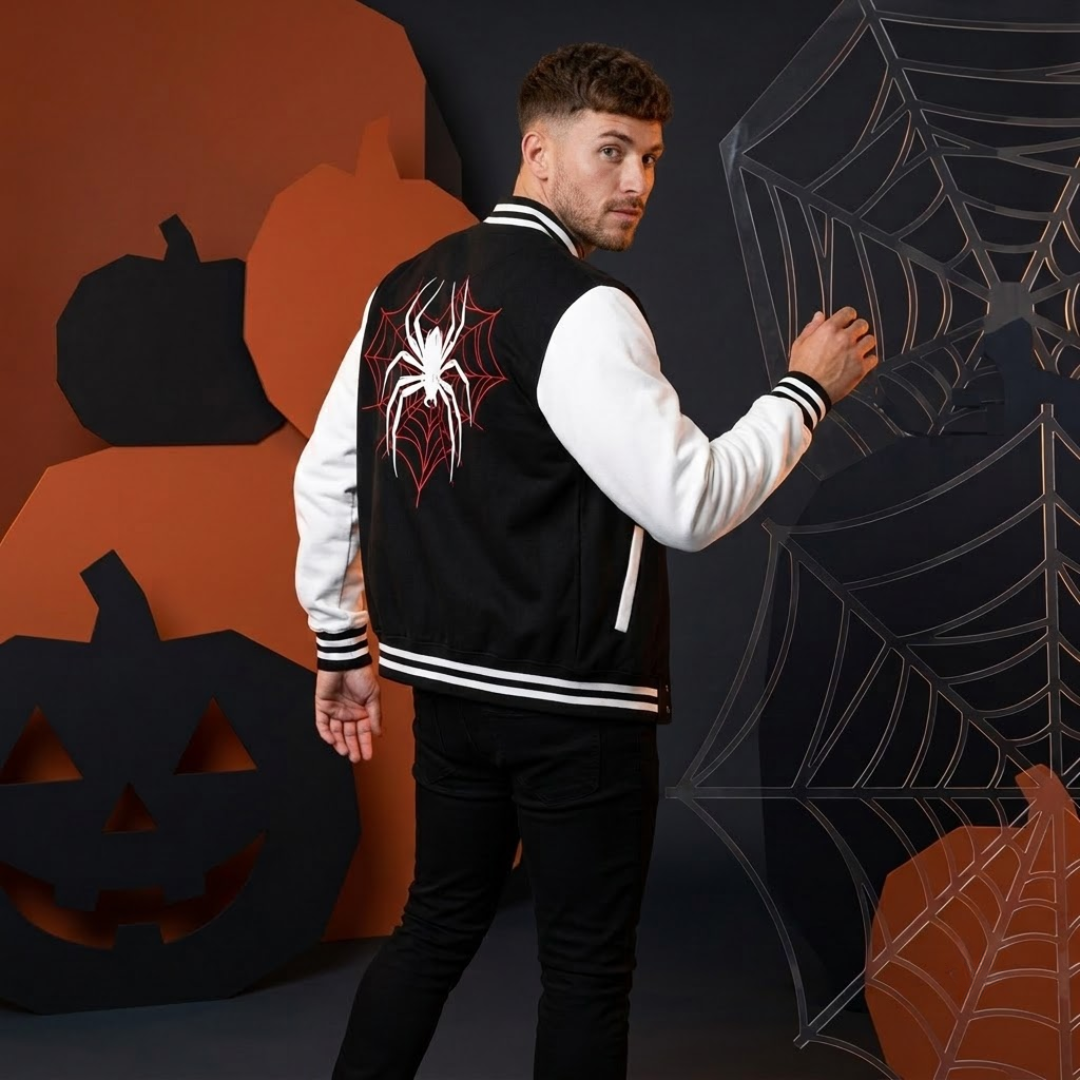 A man wearing a black and white varsity jacket with a red and white spider graphic on the back, posed against a Halloween-themed backdrop with pumpkins and spiderwebs.