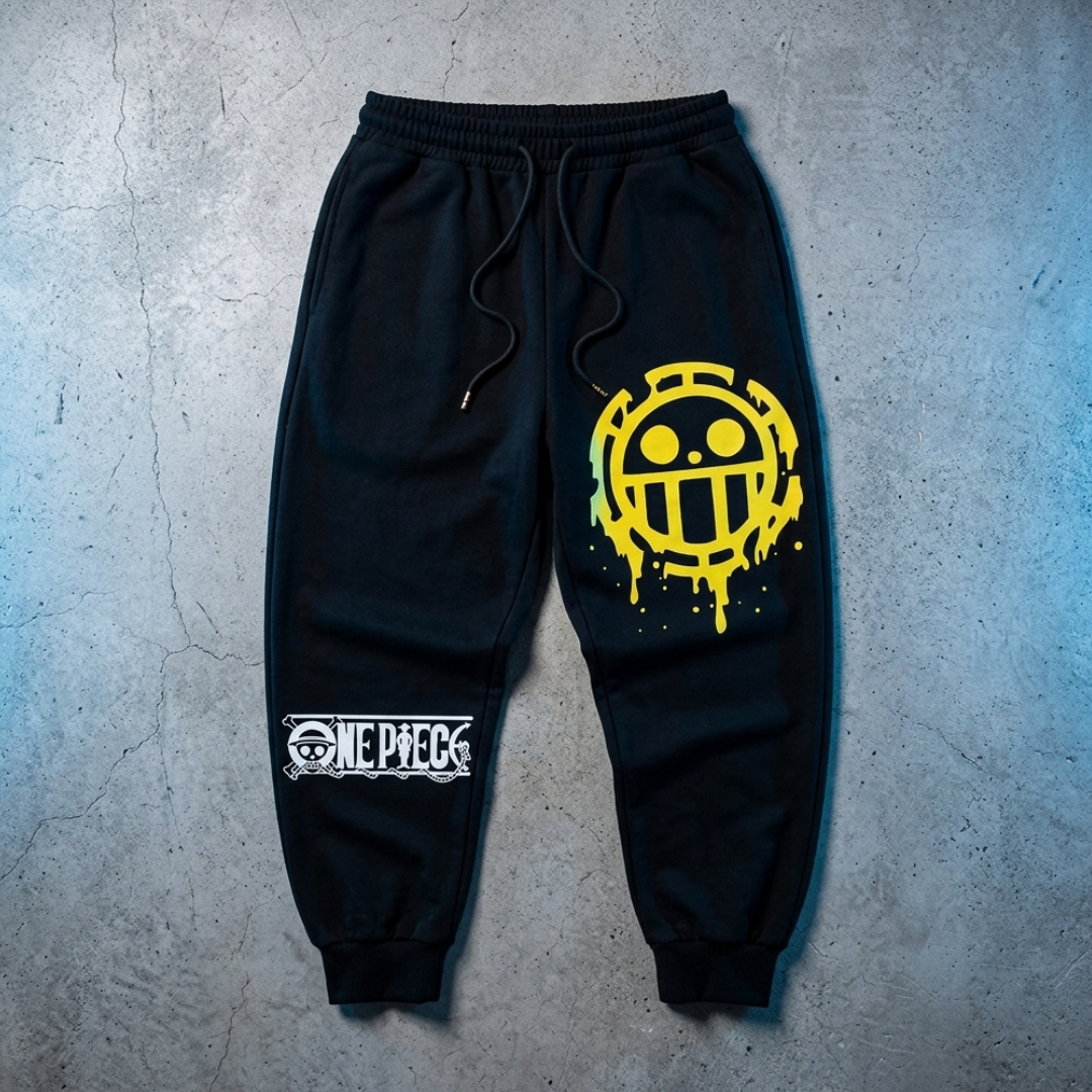 Black jogger sweatpants featuring a large yellow One Piece pirate logo on the right leg and a smaller white One Piece logo patch on the left leg, laid flat on a textured gray concrete background.