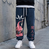 A person wearing dark navy blue sweatpants with a large pink Dragon Ball graphic on the left leg and vertical pink Japanese text on the right leg, paired with white sneakers and a black hoodie, standing against a concrete wall.