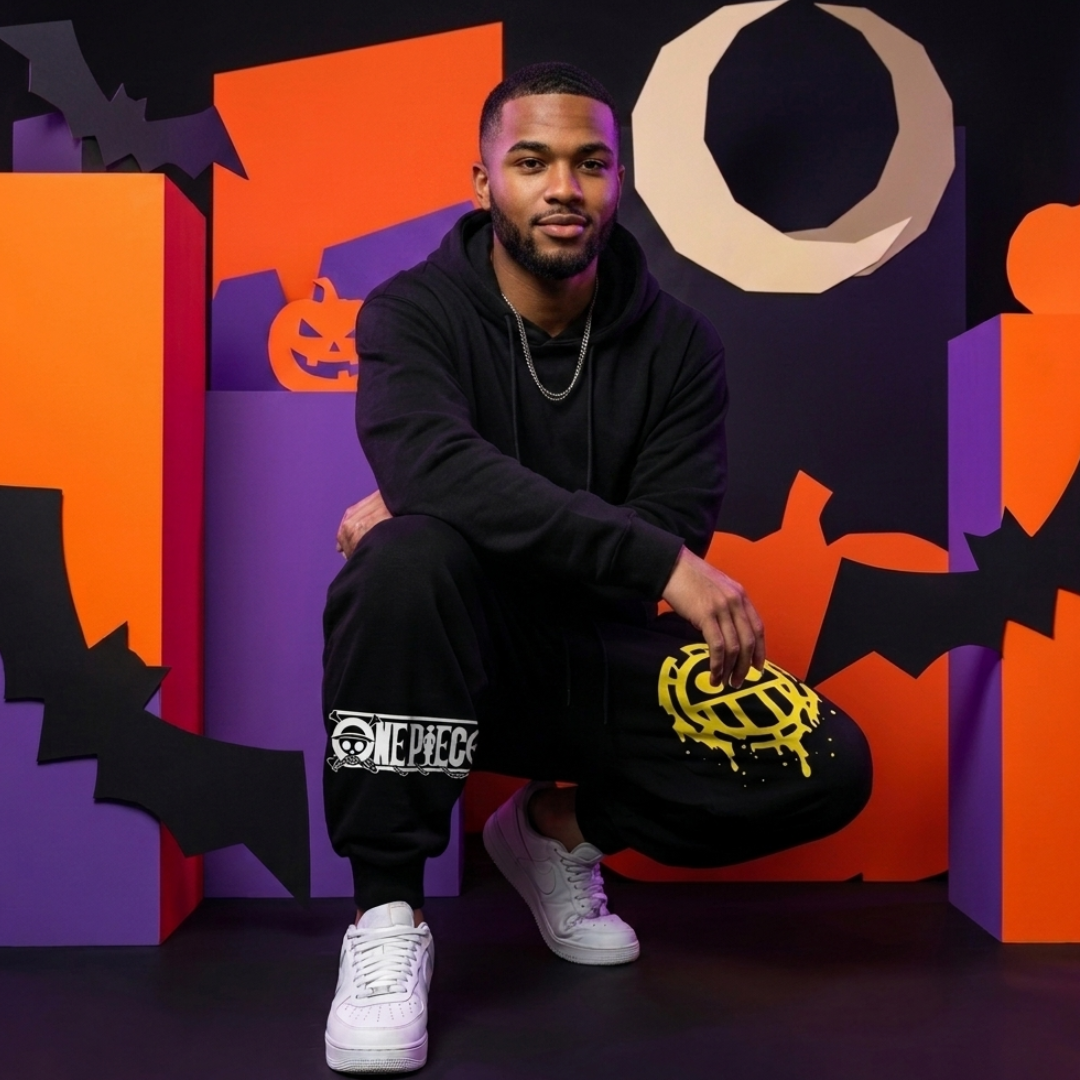 A man wearing a black hoodie and black sweatpants with One Piece graphics crouches in front of a Halloween-themed backdrop featuring orange, purple, and black shapes.
